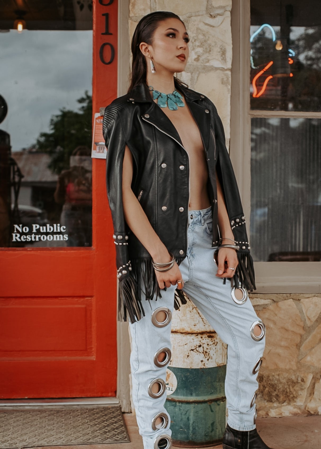 Person wearing a black leather vest with fringe, light blue jeans with button details, and black boots on a street background.